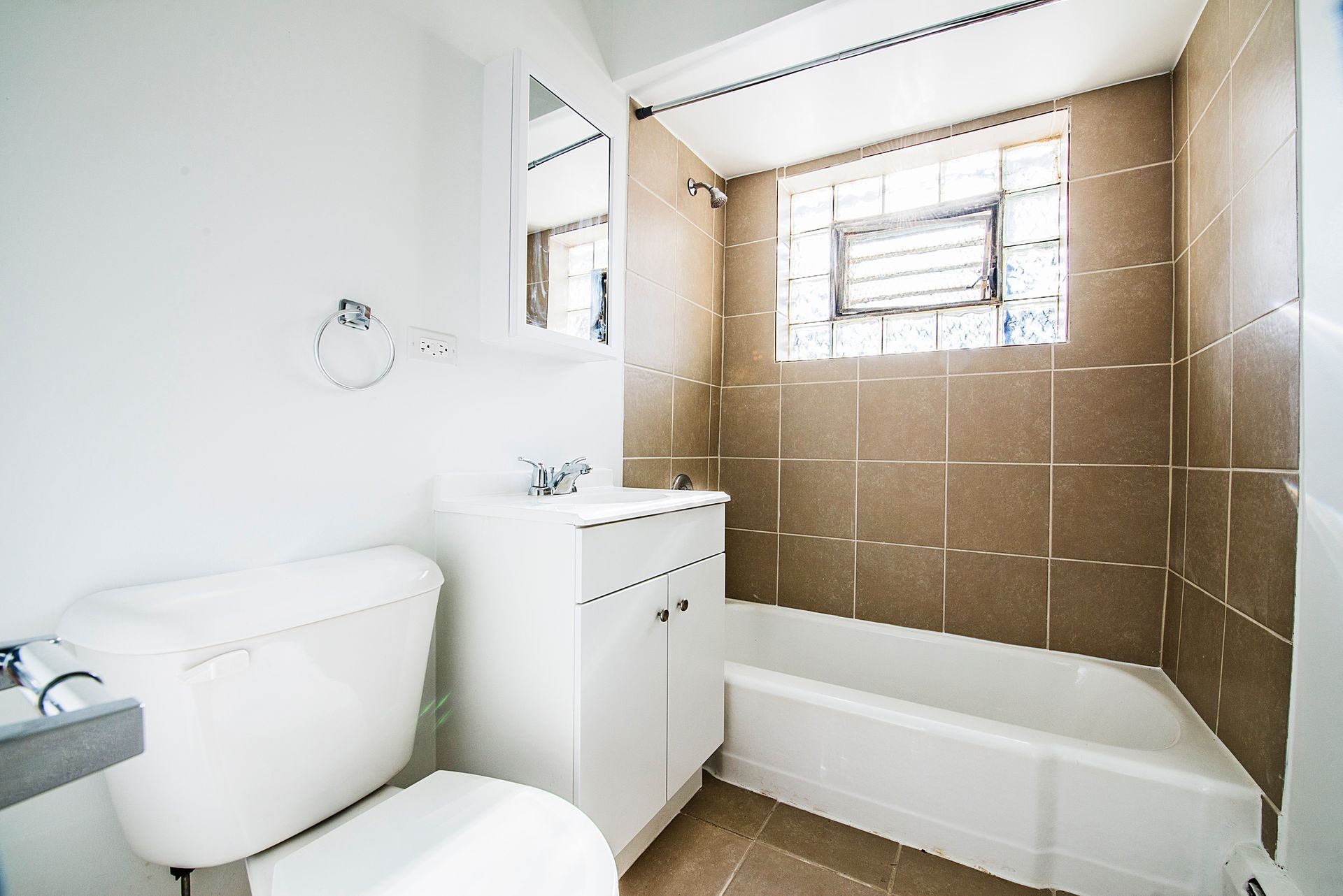 Bathroom with white toilet, vanity, and bathtub; brown tiled shower area; small window.