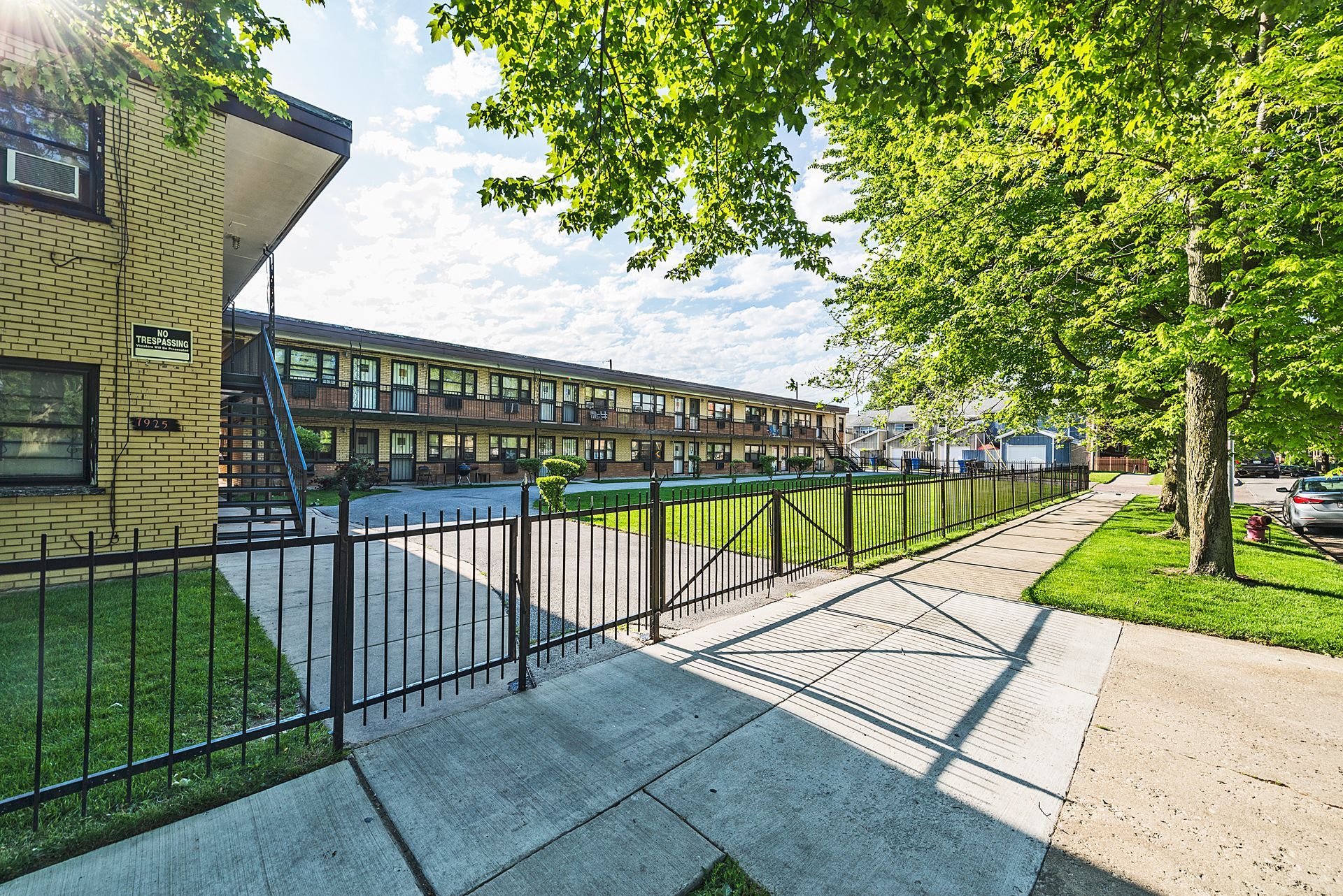 Apartment building exterior, black fence, sidewalk, green grass, trees.