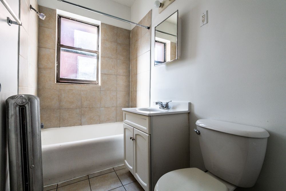 Bathroom with beige tiled walls, white tub, toilet, and vanity, and a window.