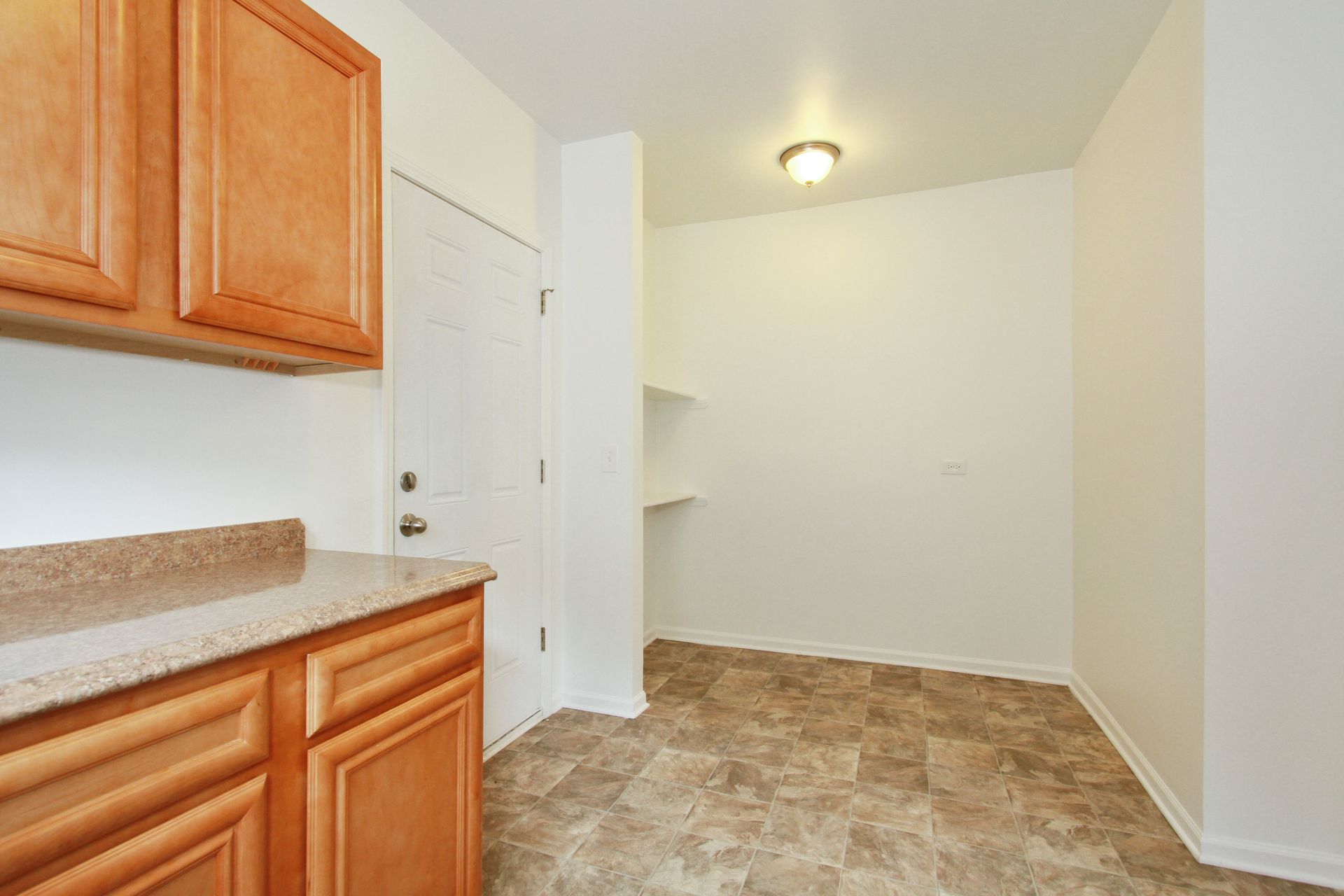 Kitchen with tan cabinets, beige countertop, door, and a shelf, beige and brown tiled floor, white walls.