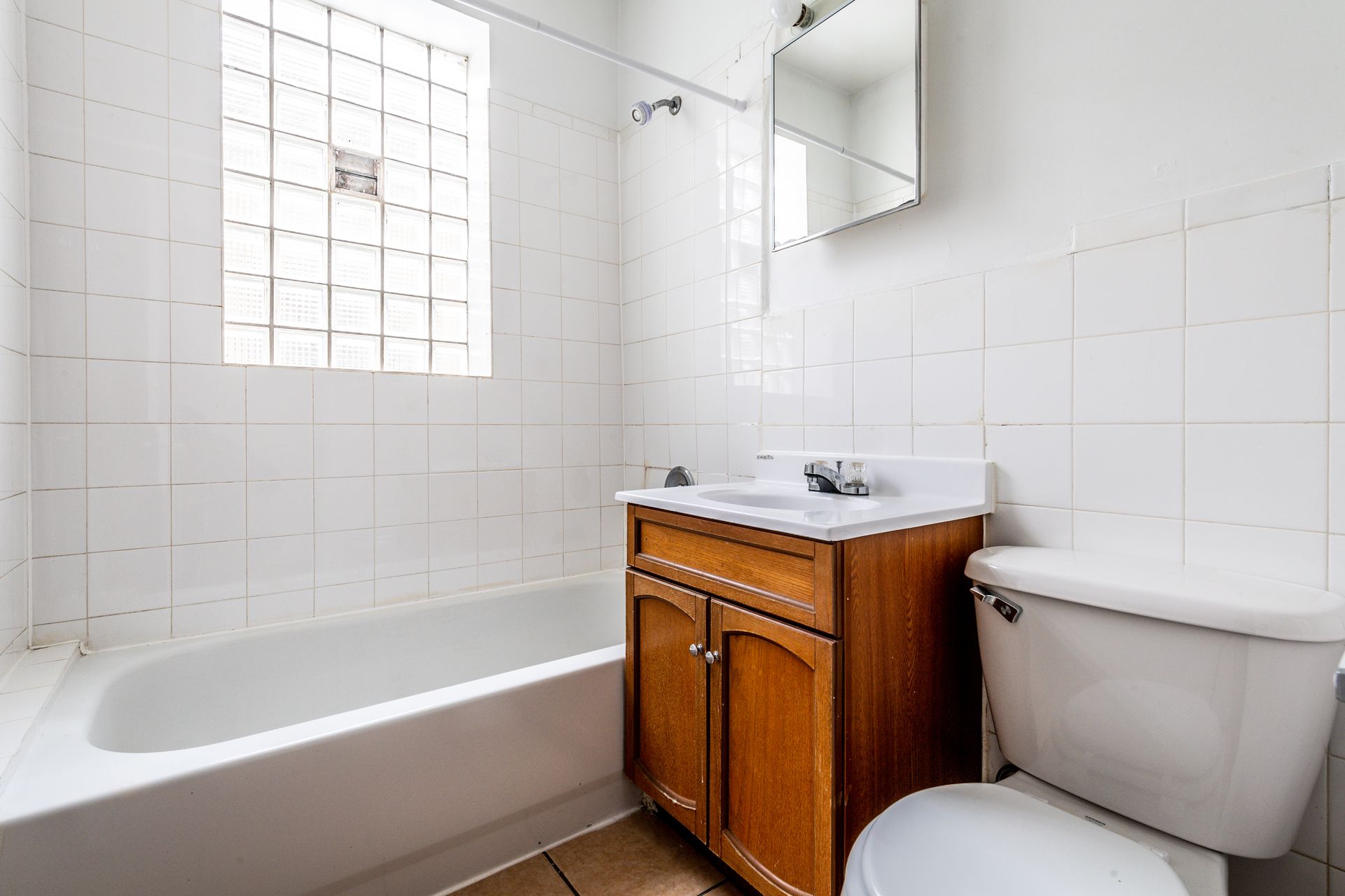 White tiled bathroom with a bathtub, vanity, toilet, and a glass block window.