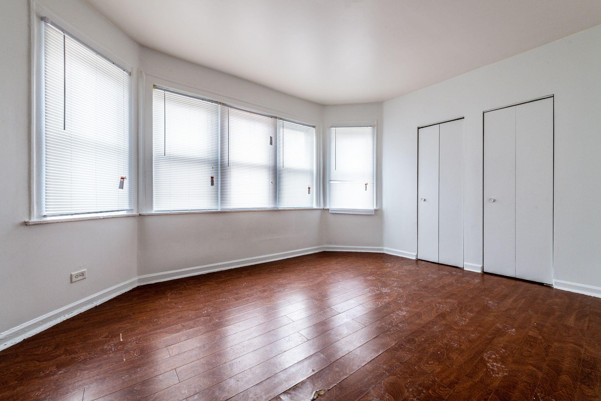 Empty room with wood floors, white walls, and a closed door. Window with blinds on the left.
