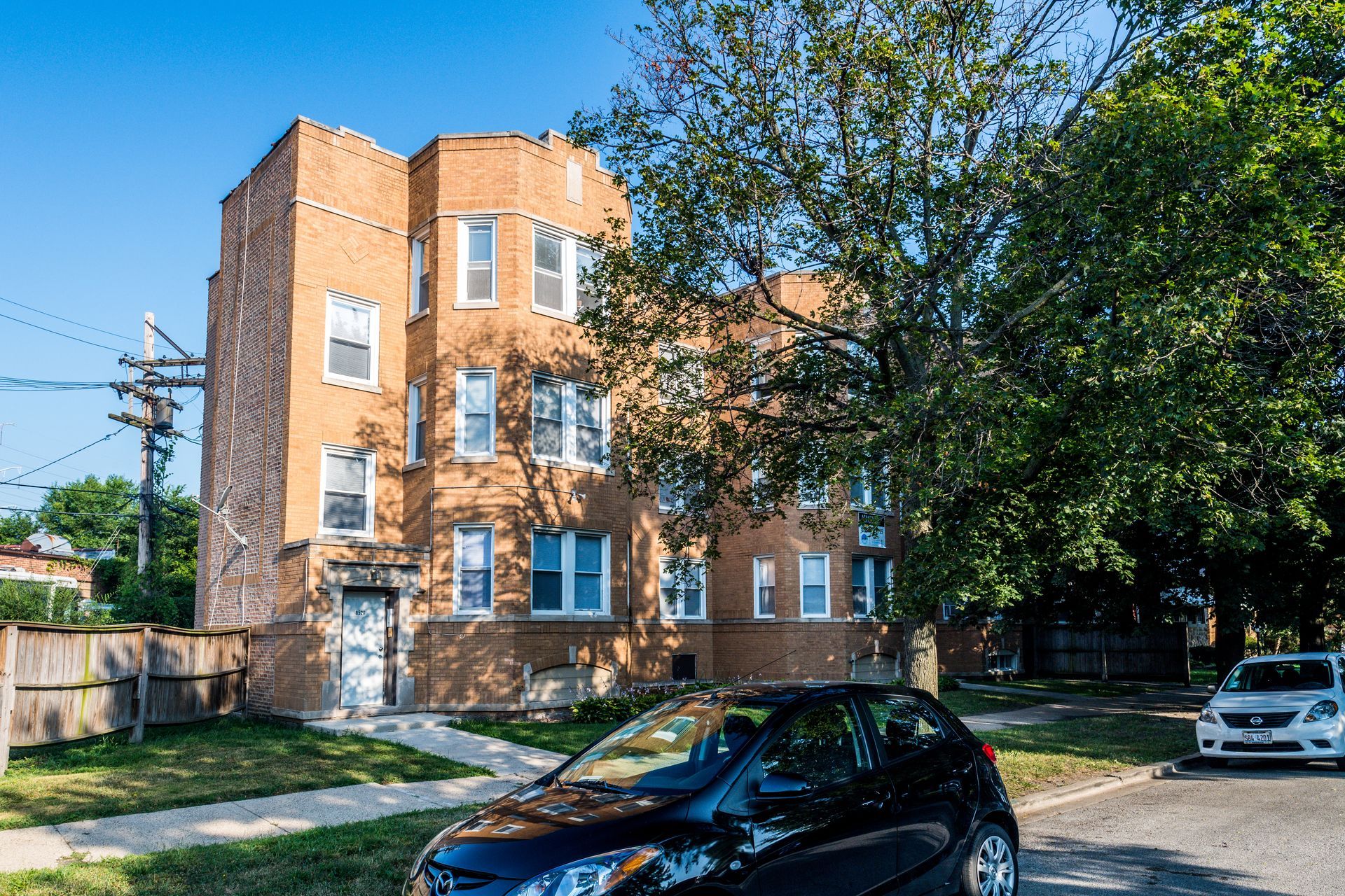 Brick apartment building with parked cars on a sunny street, tall tree in the front.