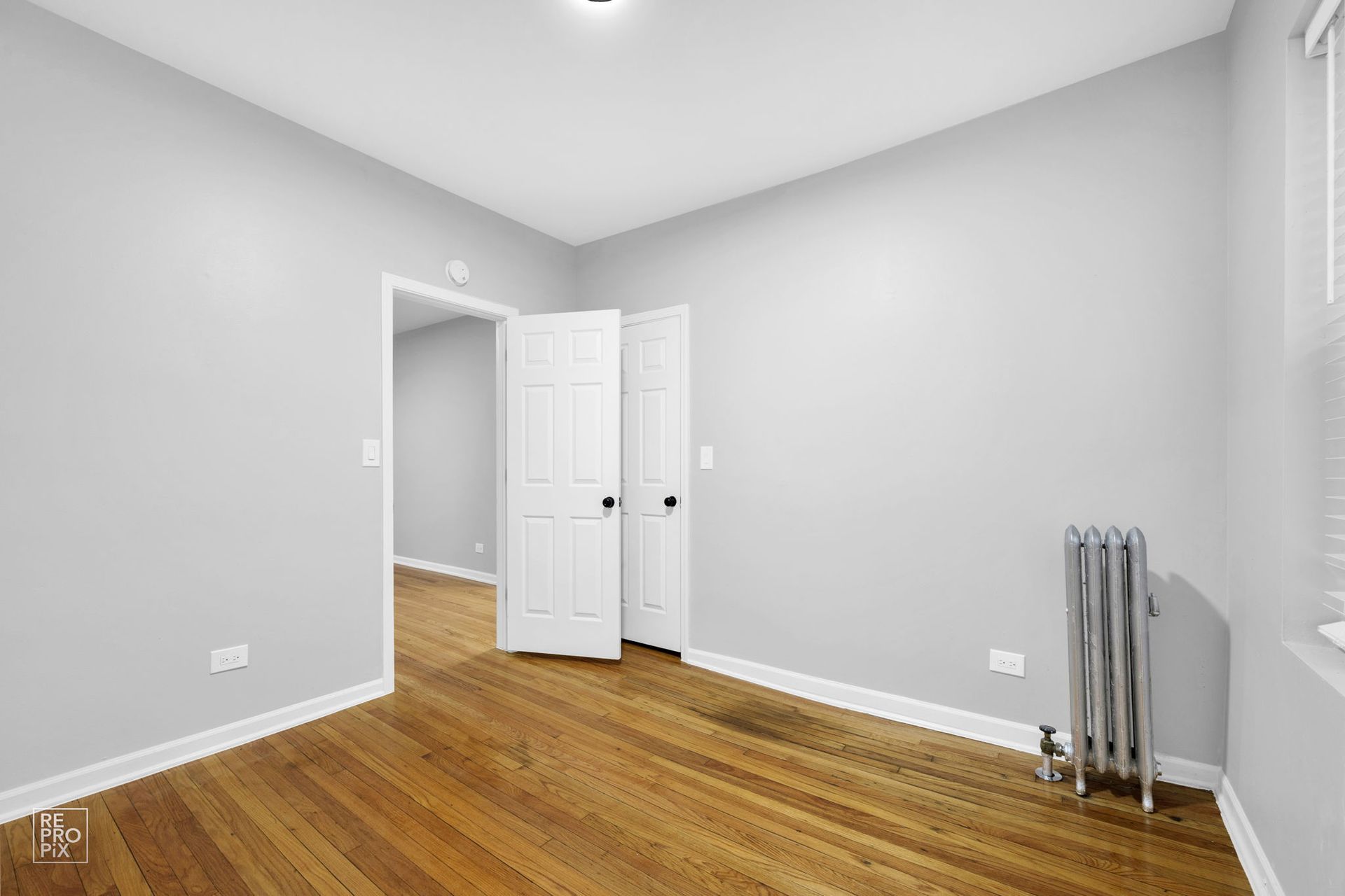 Empty room with hardwood floors, gray walls, closed white doors, and a radiator.