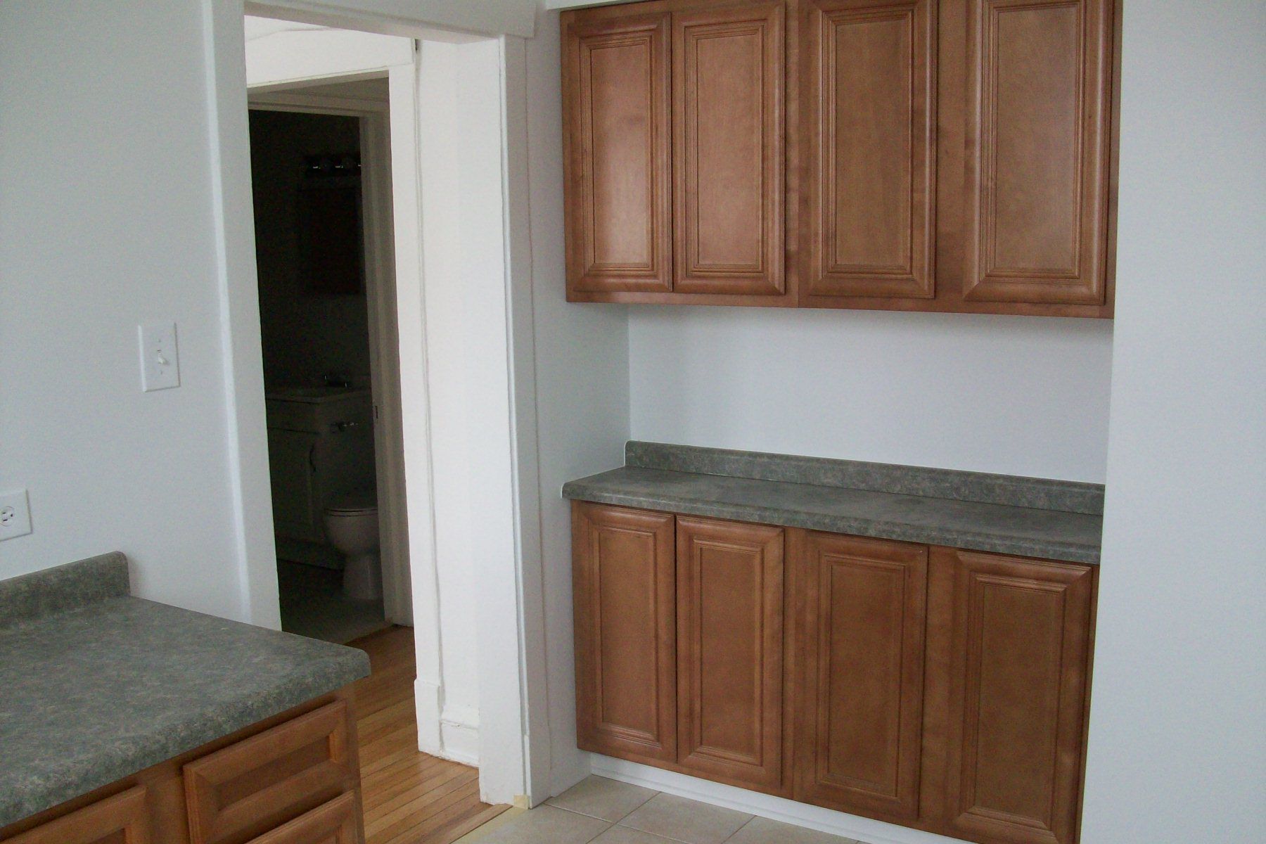 Kitchen cabinets, countertop, doorway to a bathroom. Brown cabinets, white walls, and gray countertop.