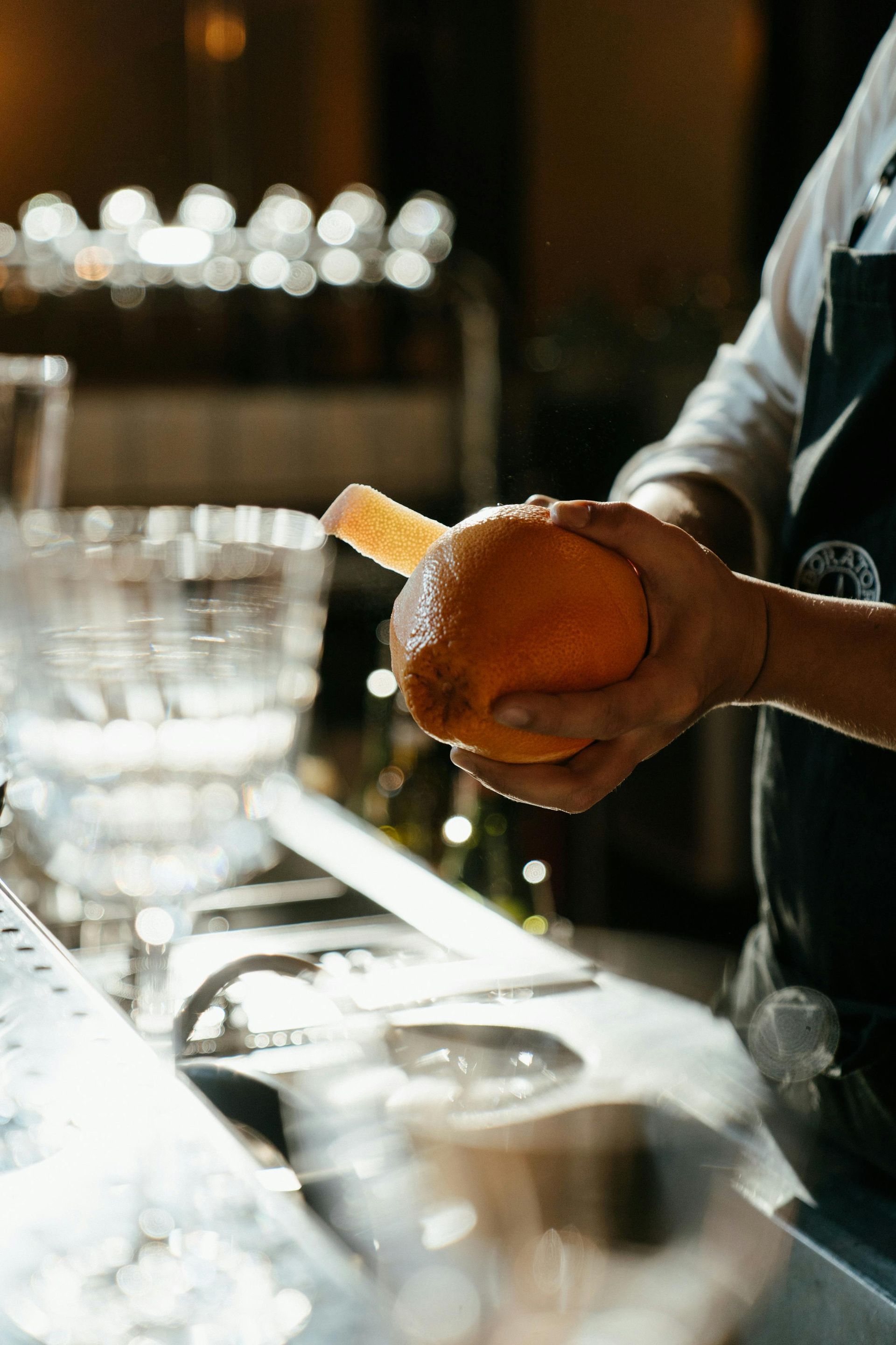 Bartender holding an orange over a lit bar counter with glassware in the background