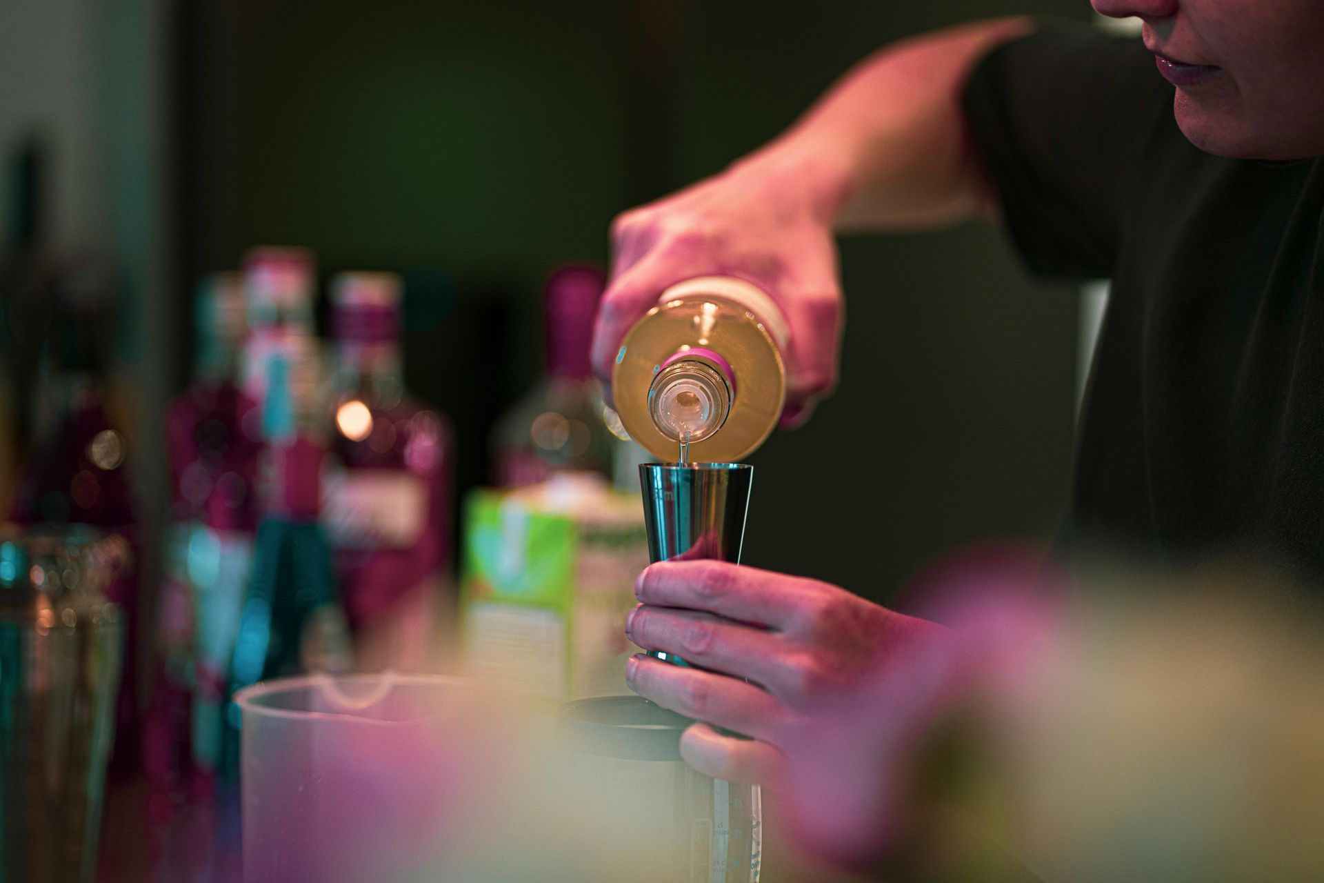 Bartender pouring a drink from a bottle into a glass at a bar, with colorful bottles blurred in the background