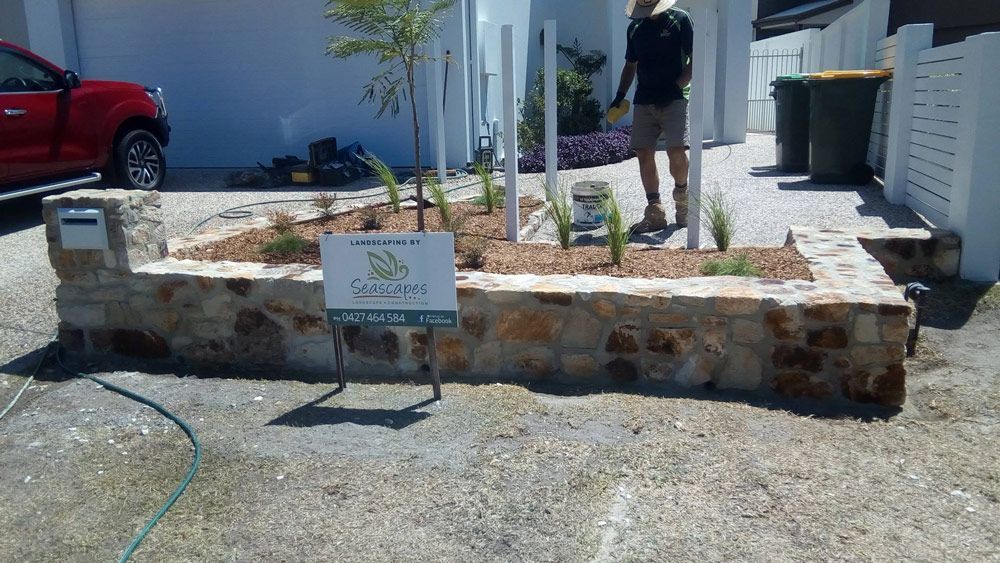 A Man is Standing in Front of a Stone Wall With a Sign That Says 'Gardens' on It — Seascapes Landscape Construction in Wauchope, NSW
