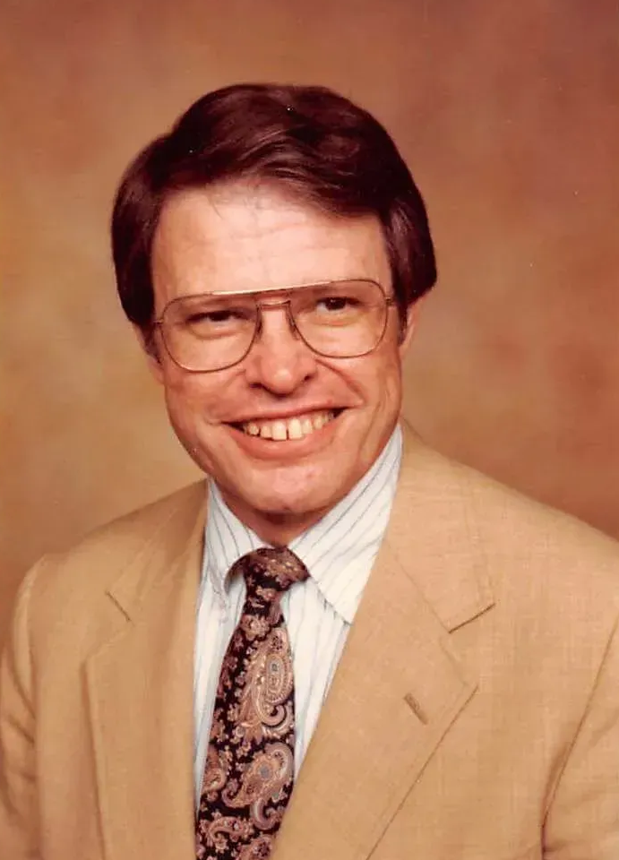 Studio portrait of a smiling man in a tan suit, striped shirt, patterned tie, and glasses on a brown backdrop