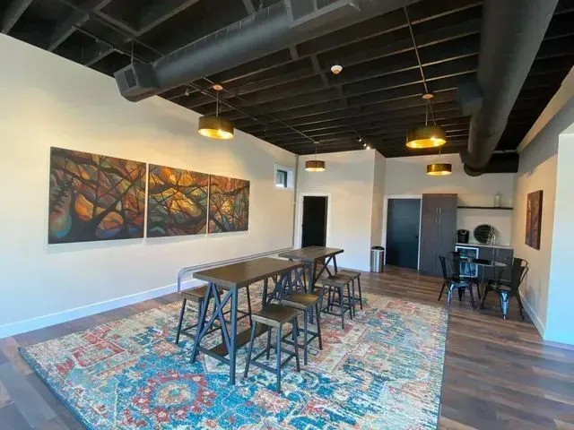 Modern lounge with high tables, stools, colorful rug, abstract wall art, and exposed ceiling beams.