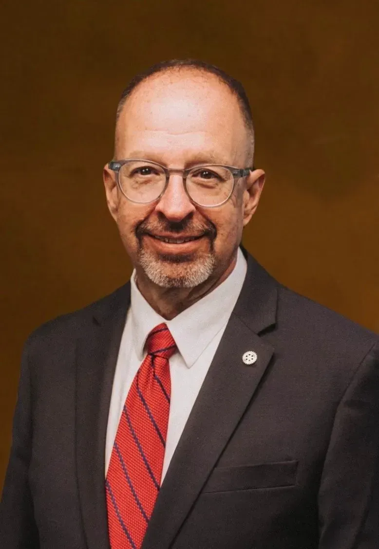 Portrait of a man in a dark suit and red tie against a brown background.