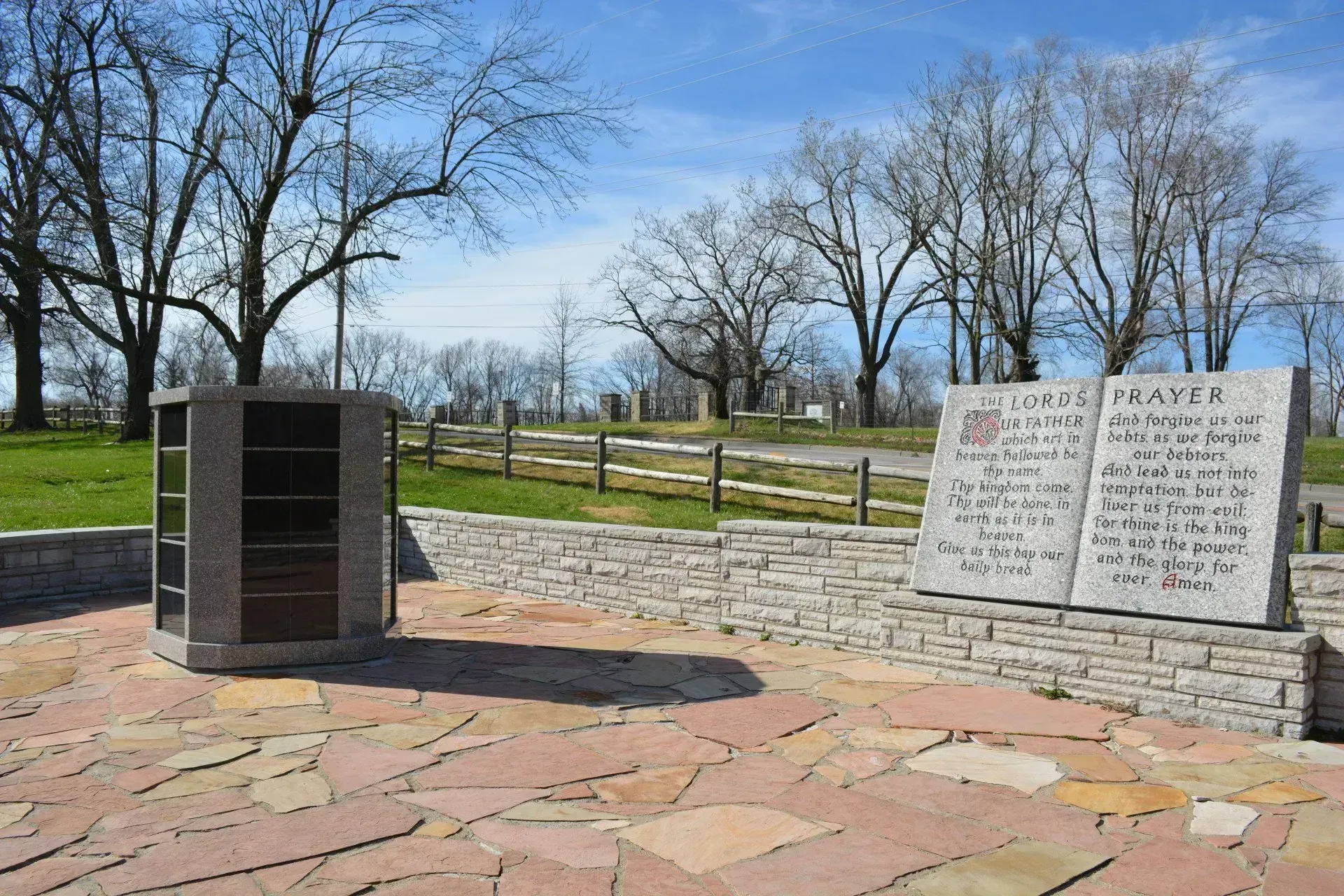Stone memorial plaques and black monument on a paved overlook in a park with leafless trees and blue sky