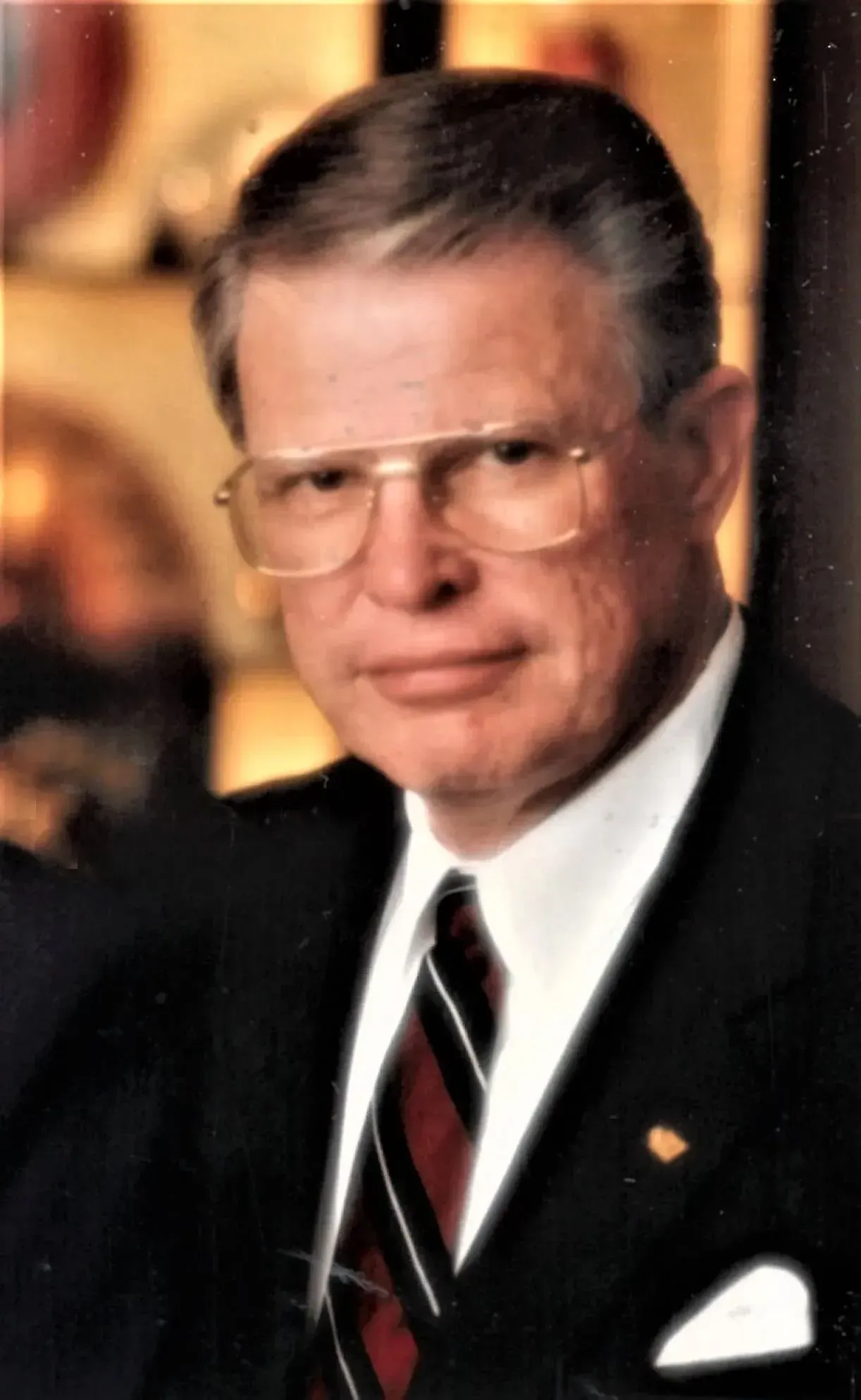 Portrait of a man in a dark suit, striped tie, and glasses against a warm blurred background