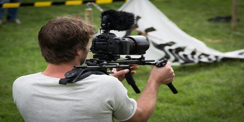 Person filming with a professional video camera on a tripod in a grassy outdoor area.