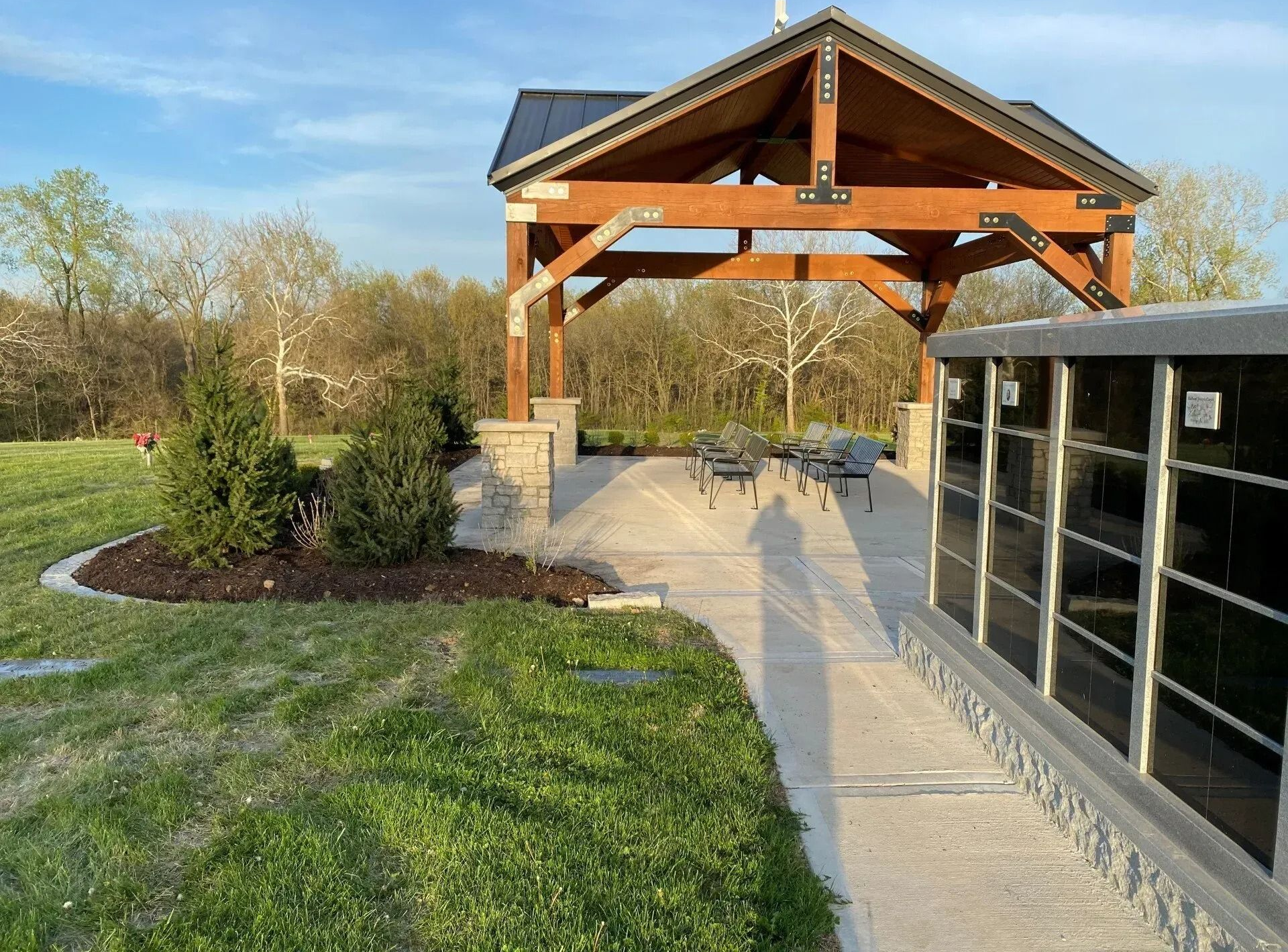 Outdoor pavilion with wooden roof, concrete walkway, grass, and a glass-walled building beside it