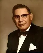 Portrait of a man in a black tuxedo, bow tie, and glasses, posed against a brown background