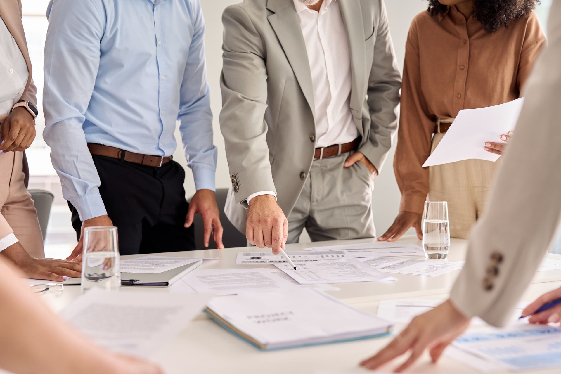 People in business attire standing around a table, reviewing documents during a meeting.