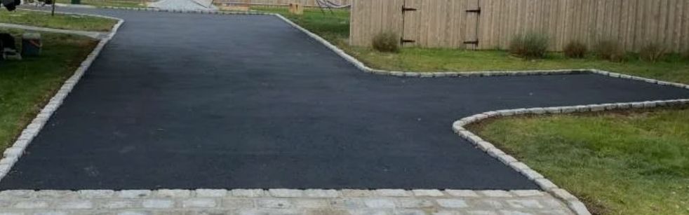 Fresh black asphalt driveway curving through a grassy yard with stone edging and a wooden fence.