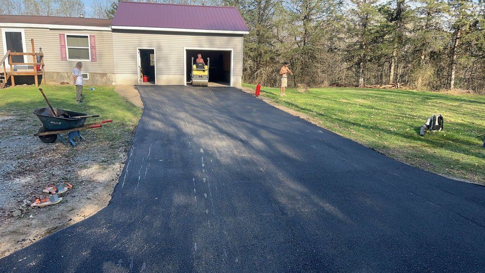 Freshly paved black driveway leading to a garage beside a house with orange cones and lawn equipment on the grass