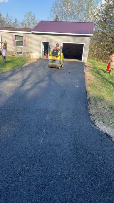 Freshly paved black driveway leading to a garage, with a worker on a yellow roller in front of the house.