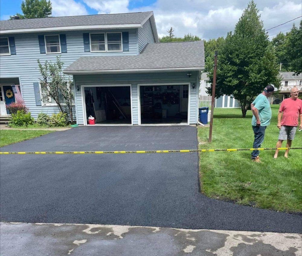 Freshly paved black driveway in front of a gray house, with two people standing on the lawn.