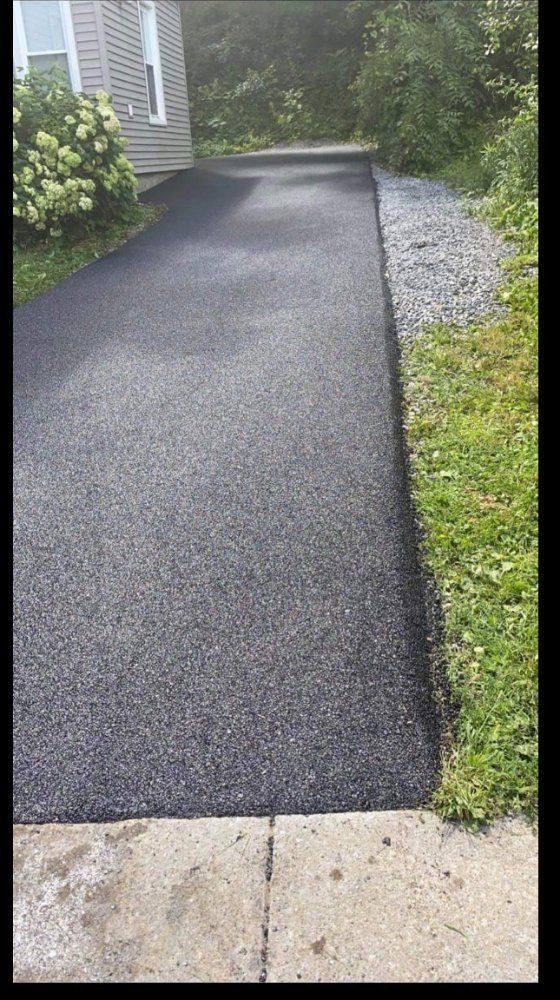 Freshly paved black walkway beside gravel and greenery, leading toward a house entrance