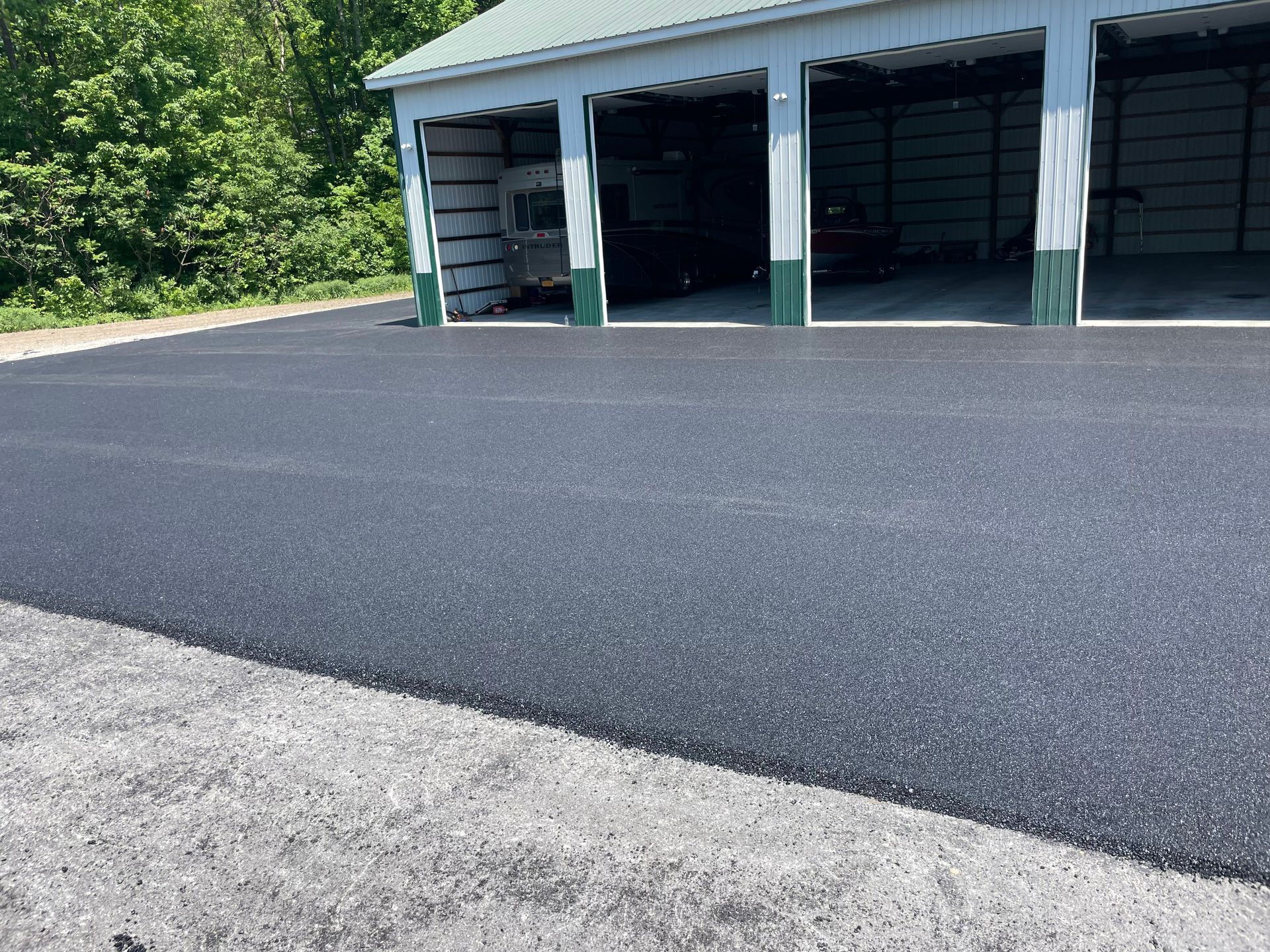 Freshly paved asphalt road in front of open garage bays with wooded area behind