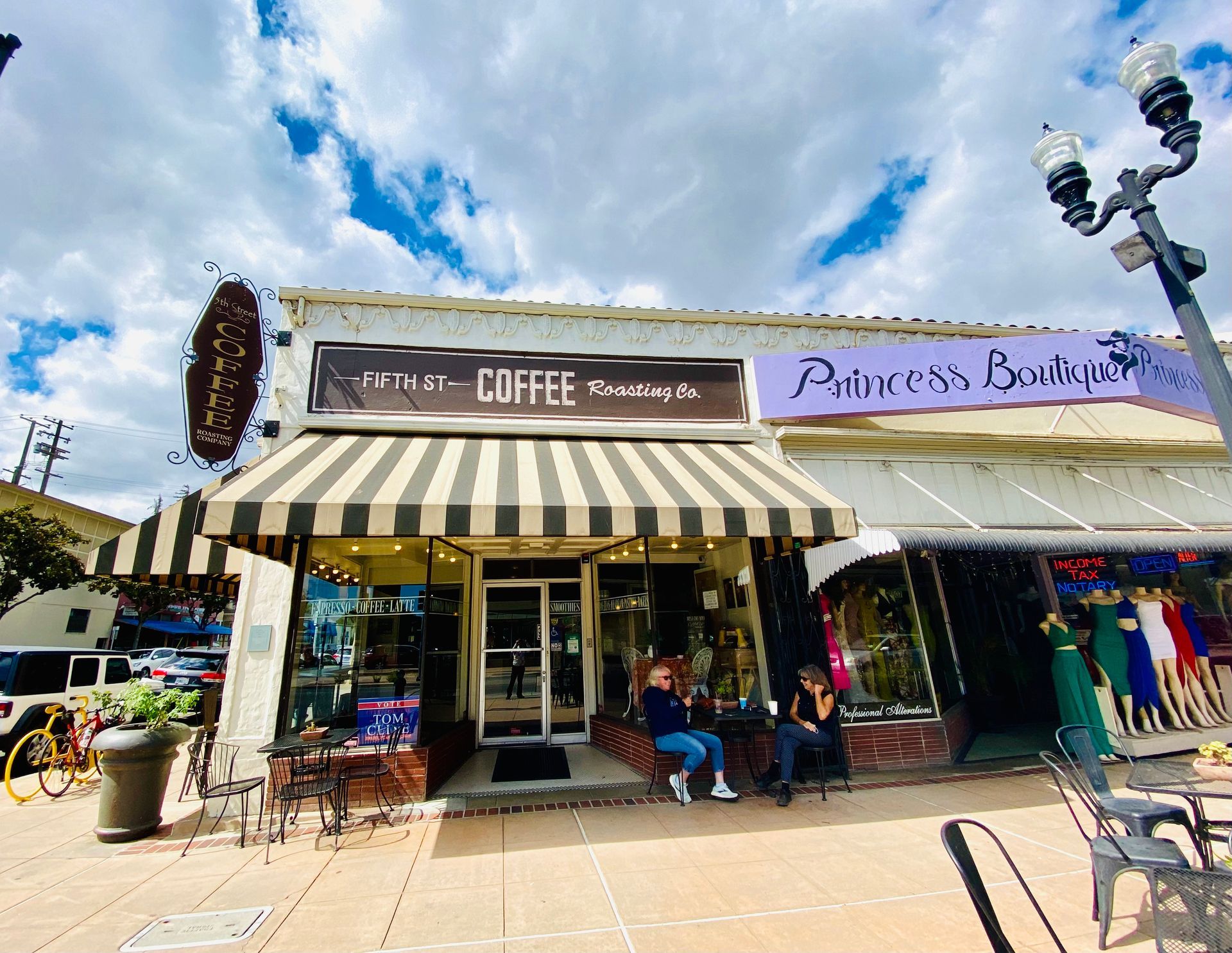 A couple of people are sitting outside of a coffee shop with a striped awning.