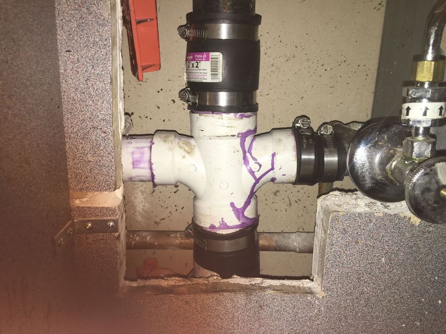 White PVC plumbing tee connected to pipes with black rubber couplings and metal clamps inside a wall cutout.