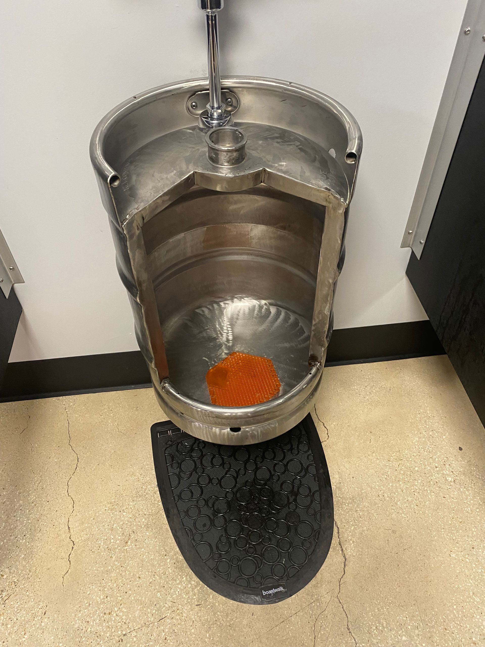 A wall-mounted urinal fashioned from a cut-open metal beer keg, featuring an orange urinal cake in a public restroom.