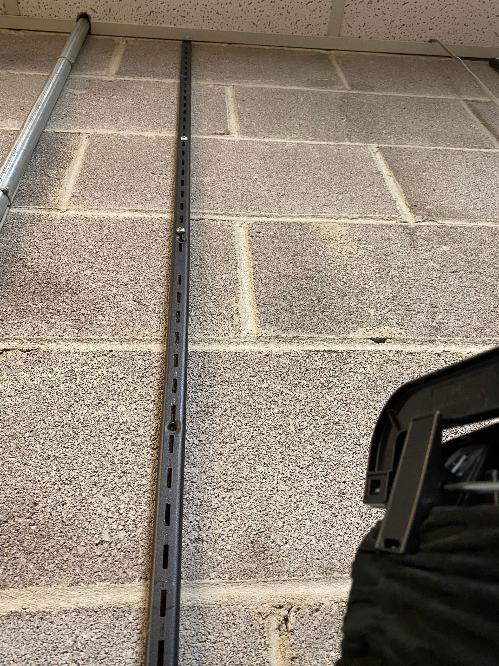 A metal wall-mounted shelving rail attached to a textured concrete block wall, next to a black hanger.