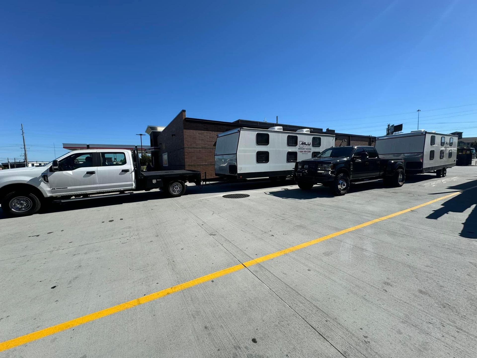 A white truck and a black truck each towing a white camper trailer parked in a paved lot under a clear blue sky.
