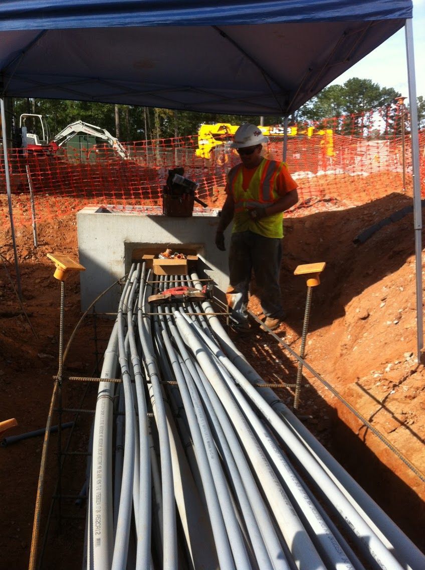 A construction worker in a hard hat and safety vest installs electrical conduits at an outdoor site under a tent.