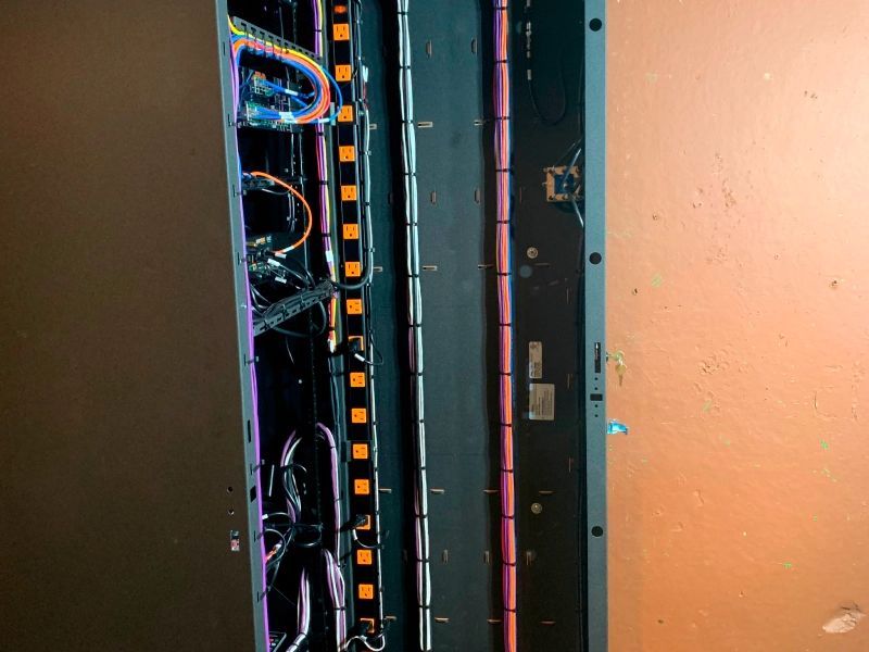 Open black utility panel with bundled, colorful wires running vertically. Orange and pink accents. Brown wall background.