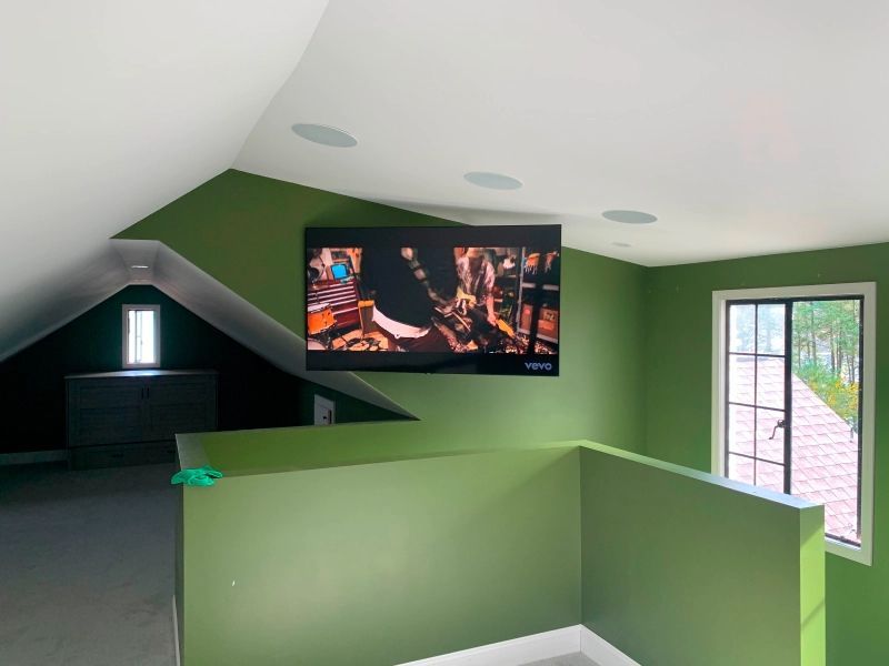 Green-painted room with mounted TV, speakers, and a partial balcony. A window and storage are in the background.