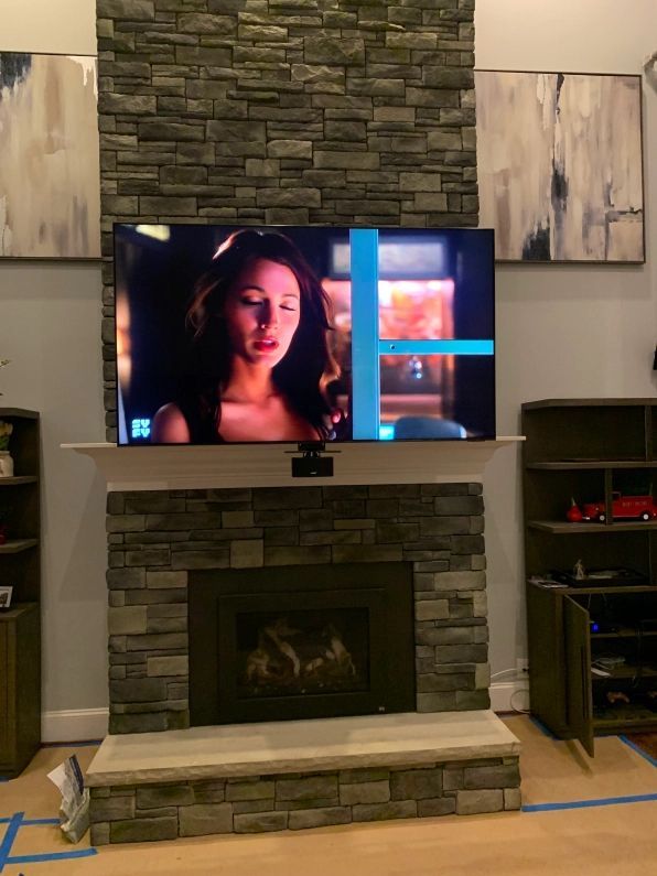 A television above a fireplace with a blue line across the screen. A woman is on the screen. Stone wall surrounds.