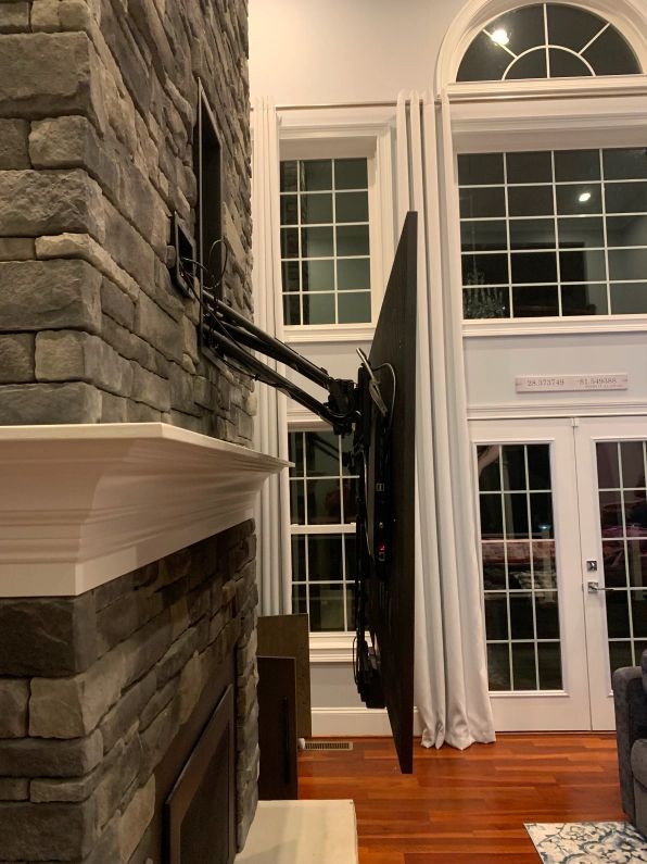 TV mounted on an articulating arm over a stone fireplace, positioned at an angle. Living room setting.