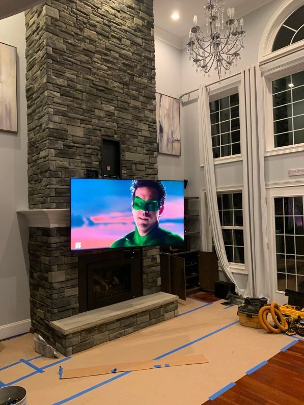 A TV playing a green-masked character is mounted on a stone fireplace in a room with large windows and a chandelier.