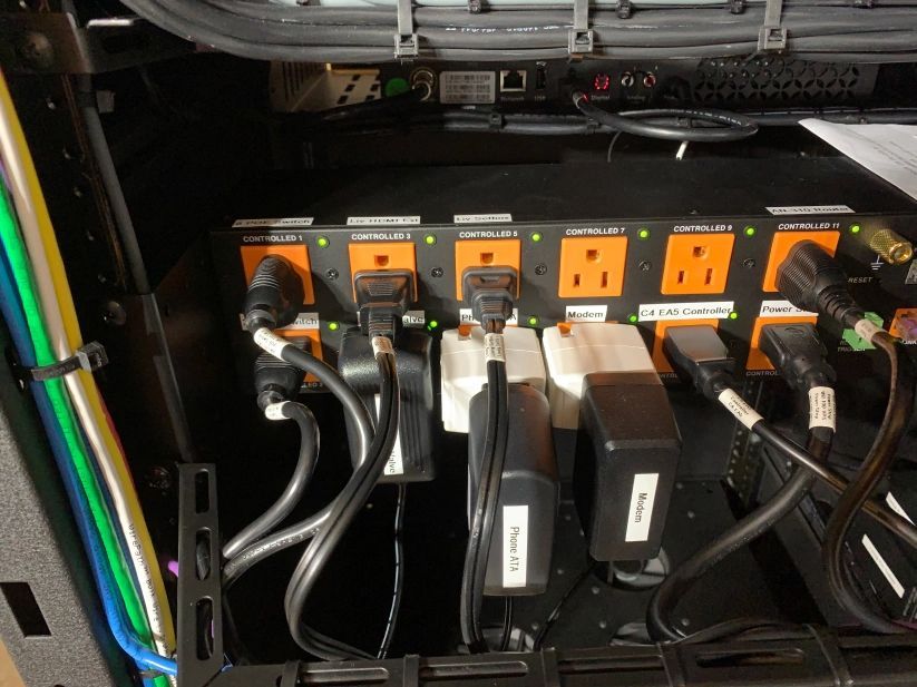 Black power strip with multiple plugs in a rack. Orange outlets and cords of various devices.