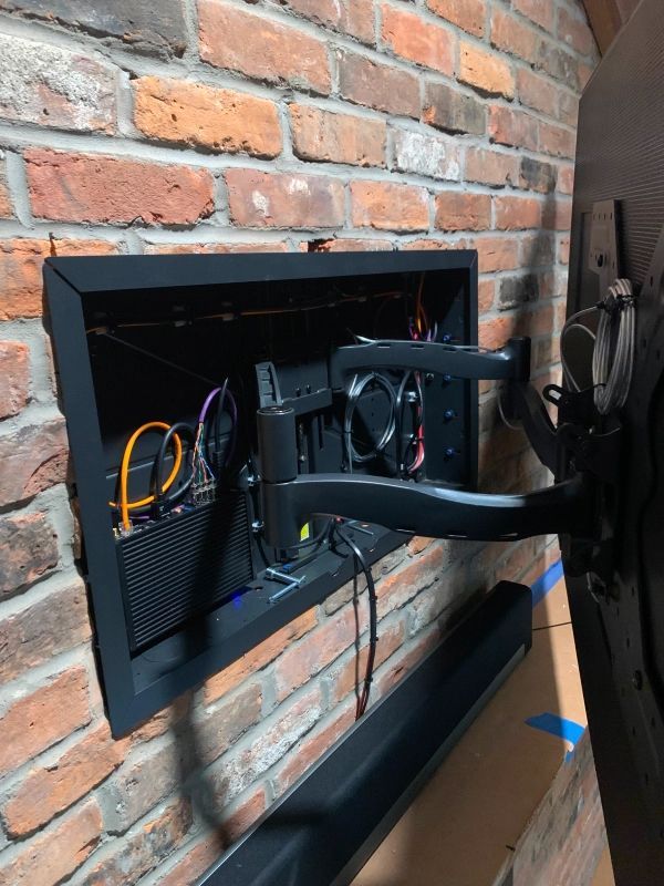 A recessed black box in a brick wall houses TV components and a mounted articulating arm. Wires and a fan are visible.