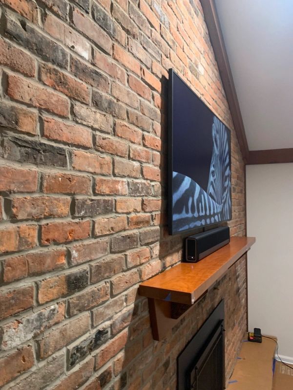 TV mounted on a brick wall above a fireplace, soundbar on a wooden shelf.