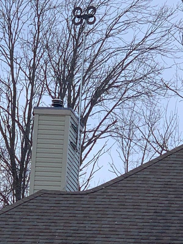 Chimney with a weather vane on top, dark roof, trees, and a cloudy sky.
