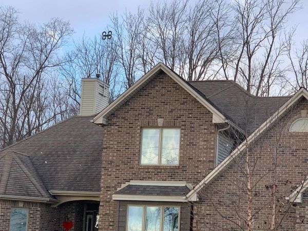 Drone hovering above a brick house with a brown roof and bare trees in the background.