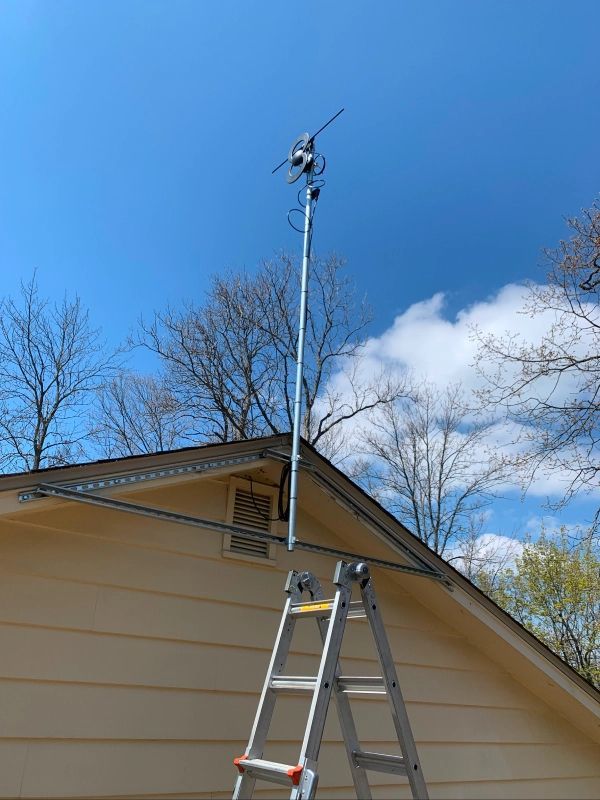 Ladder leaning against a house with a tall antenna mounted on a pole against a blue sky.