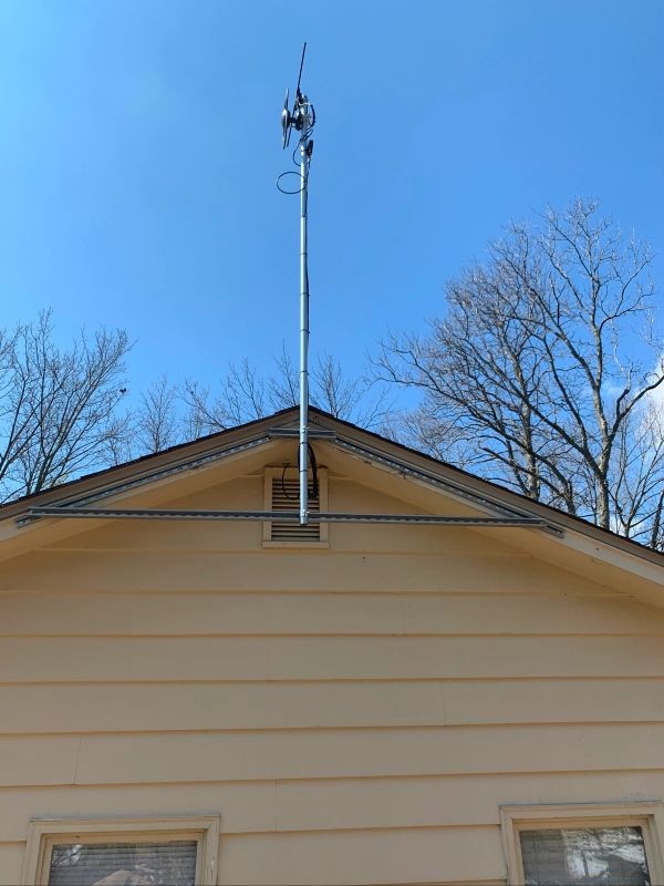 A tall antenna mounted on the roof of a yellow house against a blue sky. Bare trees are visible.