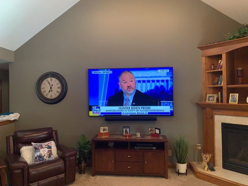 Living room with TV mounted above a cabinet. A man is on screen. Clock and fireplace nearby.