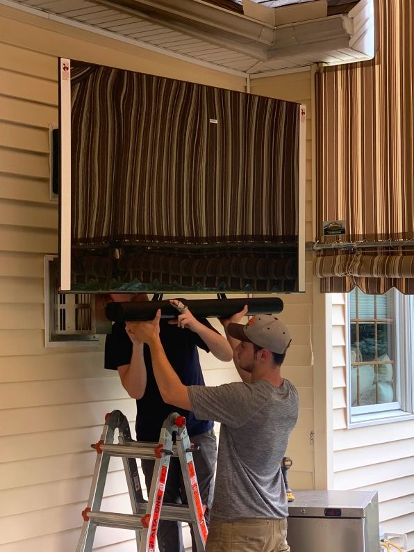 Two people mounting a large outdoor TV under an overhang. One on a ladder, holding a bracket.