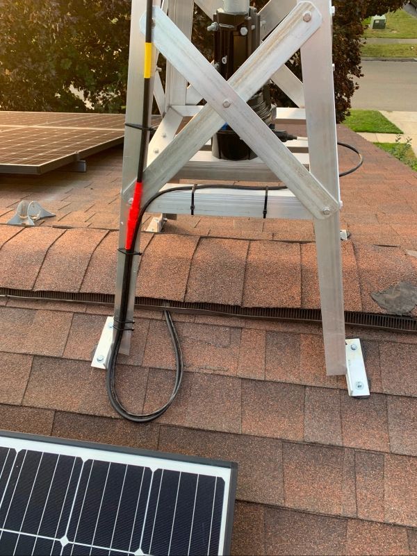 Aluminum tower on a shingle roof with solar panels. Black cable runs down the tower.