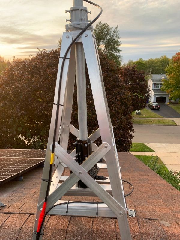 Aluminum antenna tower on a rooftop, with a cable running down to a red plug. Houses and trees in the background.