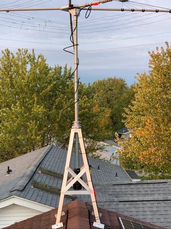 Metal antenna mounted on a rooftop with a triangular support structure, black cable, and green trees in the background.